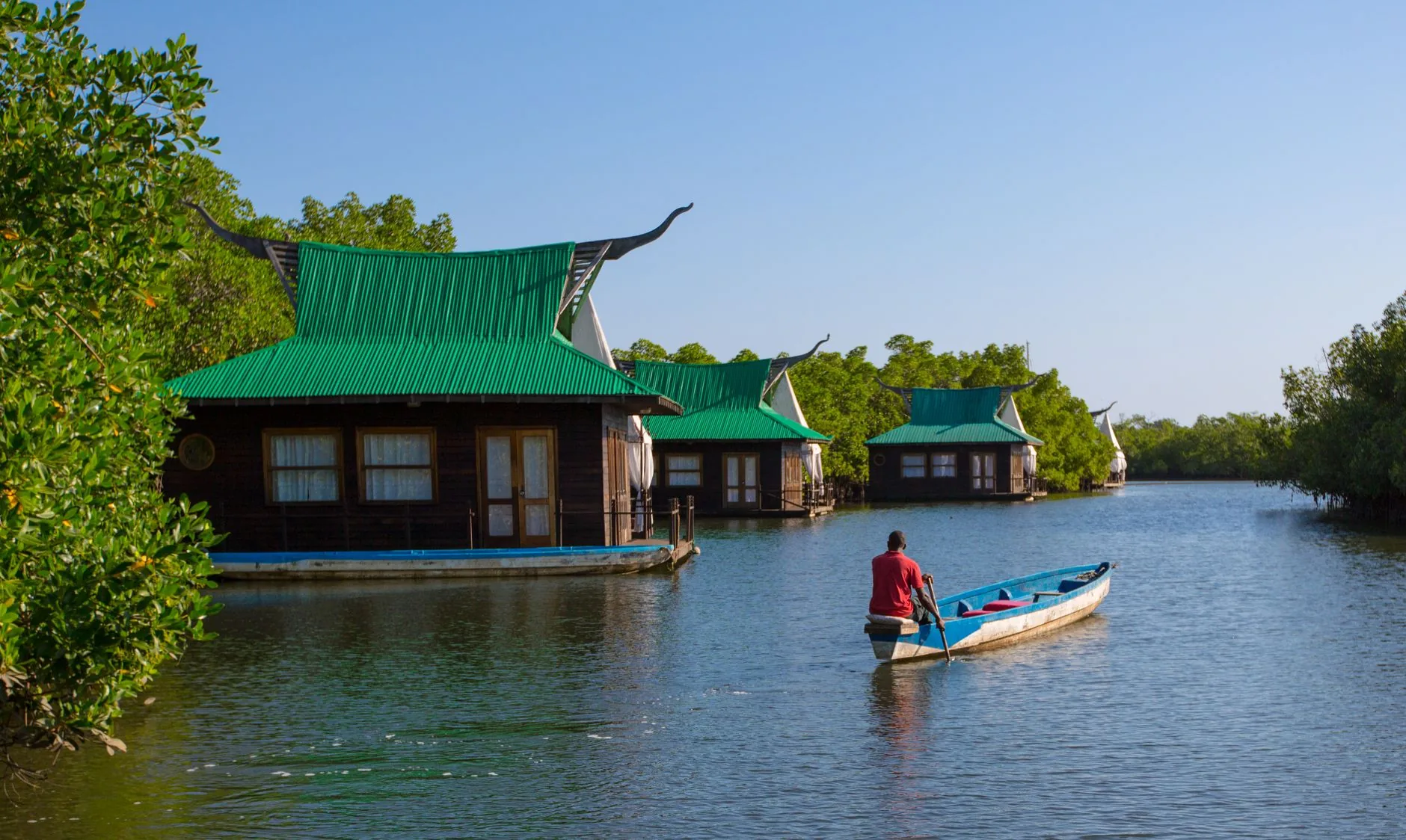 Un homme pagaie une pirogue bleue vers des lodges flottants sur une rivière bordée de mangroves